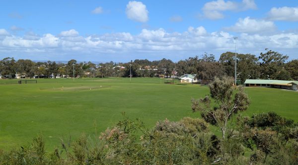 Large irrigated sports fields and open turf areas in a community recreation precinct, bordered by trees and facilities under a partly cloudy sky.