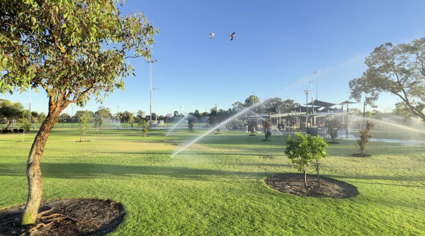 park-irrigation Automated irrigation sprinklers watering a landscaped public park with turf, young trees, and a community recreation area under a clear blue sky.